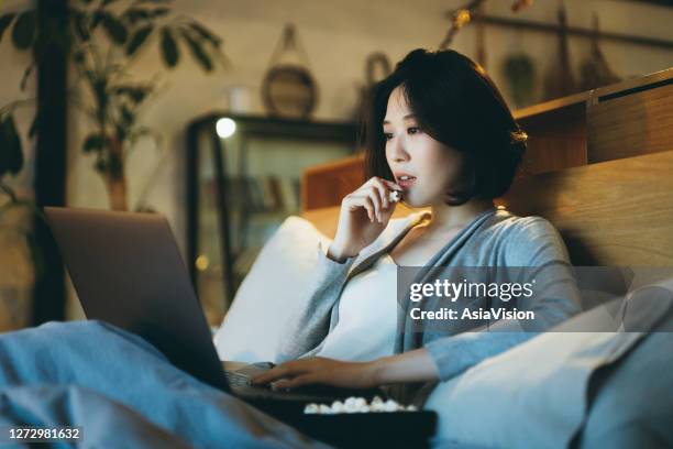 junge asiatische frau auf dem bett liegen deiner freude am wochenende, film am laptop und abends popcorn zu hause essen - asian cinema stock-fotos und bilder