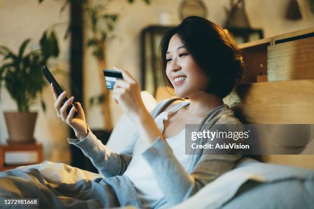 beautiful smiling young asian woman lying on the bed, shopping online on her smartphone and making mobile payment with credit card at home in the evening - online bank service stock pictures, royalty-free photos & images