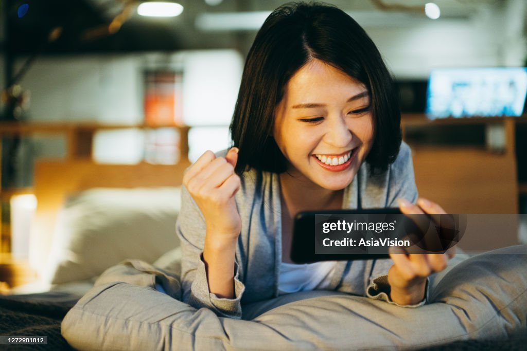 Aufgeregt junge asiatische Frau liegt auf dem Bett im Schlafzimmer, spielen mobiles Spiel auf dem Smartphone am Abend zu Hause