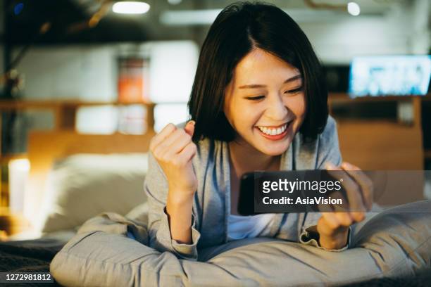 giovane donna asiatica eccitata sdraiata sul letto in camera da letto, che gioca a gioco mobile su smartphone la sera a casa - eccitazione foto e immagini stock