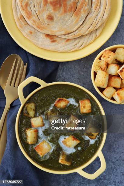 bild von behandeltem, gelbem serviergericht, vegetarischemahlzeit palak paneer (hüttenkäse, spinatpüree) rezept neben einem blau-grauen käsetuch, goldlöffel und gabel, lachha paratha (fladenbrot) und schüssel mit gewürfelten quark, blau-grauer hinte - parantha stock-fotos und bilder