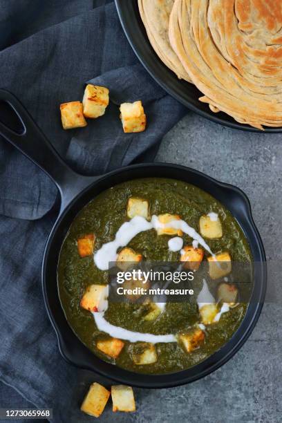 image of cast iron round skillet frying pan, vegetarian meal palak paneer (cottage cheese, spinach puree) recipe, blue-grey cheese cloth, lachha paratha (flatbread), scattering of fried curd cheese cubes on blue-grey background, elevated view - palak paneer stock pictures, royalty-free photos & images