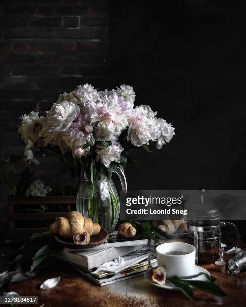 food photography of still life side view with a bouquet of peonies flowers on a breakfast table with a tea mug, croissants, books and magazines against a brick wall and a chalk board for writing text - table brick wall wood stock-fotos und bilder