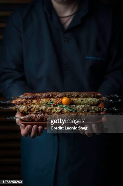 food photography of caucasian male chef front view in a blue coat holding a brown ceramic dish with kebab from different types of meat with a decoration of vegetables - chef coat stock pictures, royalty-free photos & images