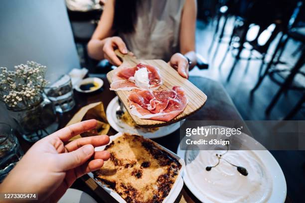 high angle view of young asian woman passing on a plate of prosciutto platter during meal over a table. enjoying a sumptuous italian meal on a date night - asian couple dining stockfoto's en -beelden