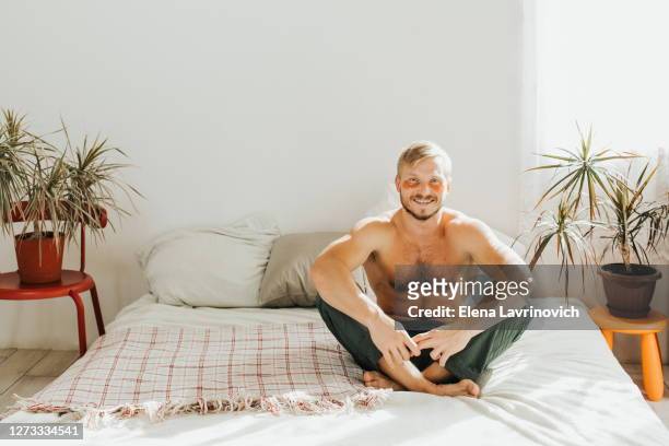 happy man is sitting on his bed at home. cosmetic patches on the face, care for the skin around the eyes. - mask man stock pictures, royalty-free photos & images