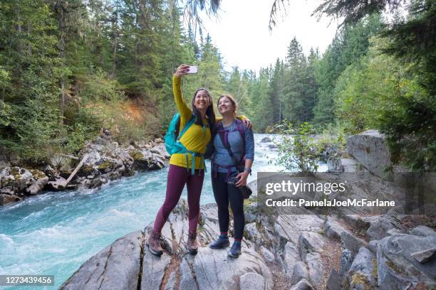 hiking sisters taking selfie on rocky ledge near river - sisterhood stock pictures, royalty-free photos & images