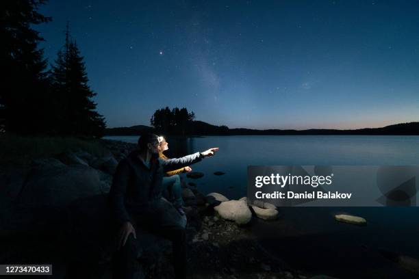 reaching for the stars together. wide shot of a couple tourists in love standing on a rock at dusk and looking at the majestic starry sky over a beautiful mountain lake. inspiration and spirituality. mental health. - reach stars stock pictures, royalty-free photos & images