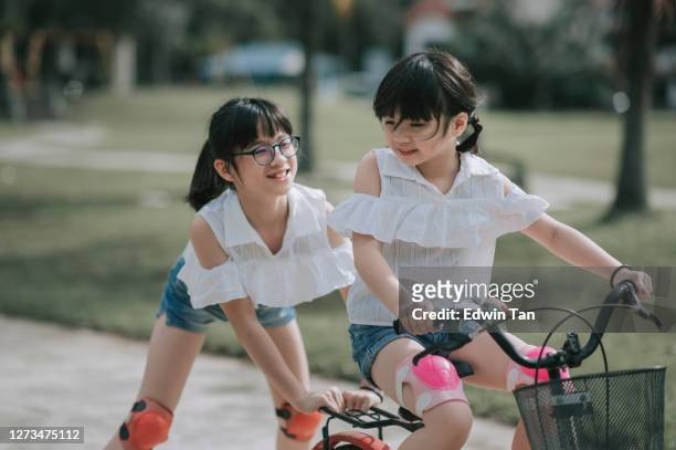 asian chinese sister pushing and helping her sister learning riding bicycle at public park in weekend morning - two kids with cycle imagens e fotografias de stock