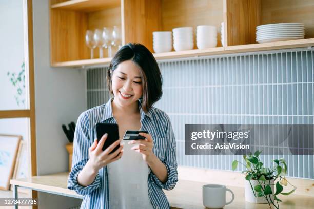 beautiful smiling young asian woman managing financial bill payment with smartphone and credit card in the kitchen at home. technology makes life so much easier - asian credit card imagens e fotografias de stock