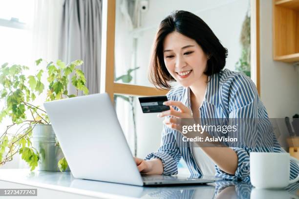 beautiful smiling young asian woman managing online banking with her laptop and credit card over the kitchen counter at home. technology makes life so much easier - asian credit card imagens e fotografias de stock