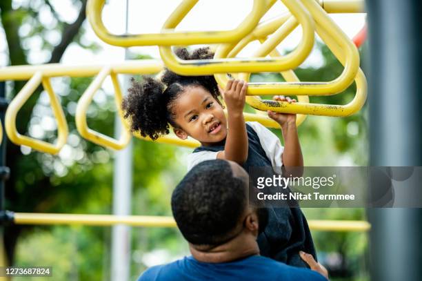 african father supporting his curly daughter while learning to play the monkey bars in jungle gym. family learning outdoors together. - cage à poules photos et images de collection