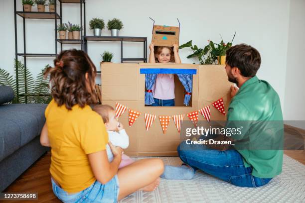 young two generation family at home, watching daughter on the stage behind cardboard box - puppet theatre stock pictures, royalty-free photos & images
