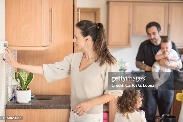 young mother adjusts the temperature of her household on a thermostat while her partner holds their baby boy in his arms  and and their daughter walks around the the kitchen - kitchen straighten stockfoto's en -beelden
