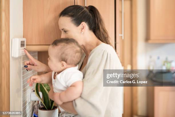 young mother holds her baby boy on her arms and shows him how to adjusts the temperature of the household on a thermostat in the kitchen - panel stockfoto's en -beelden