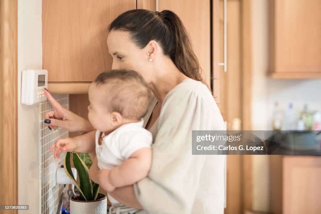 Young Mother Holds Her Baby Boy On Her Arms And Shows Him How To Adjusts  The Temperature Of The Household On A Thermostat In The Kitchen High-Res  Stock Photo - Getty Images