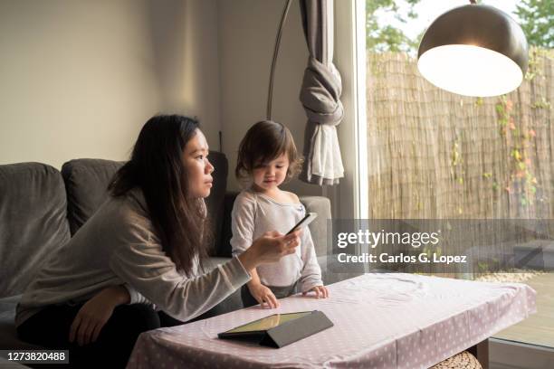 a young asian mother uses her phone to control the lights in her living room and looks up at a lamp in the living room as her daughter stands beside her - reportage home stock pictures, royalty-free photos & images