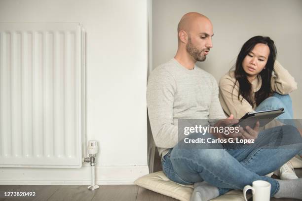a young couple sit on the floor learning how to control the heating from his digital tablet in the living room - reportage home stock pictures, royalty-free photos & images
