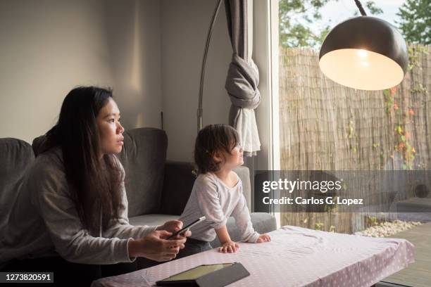 a young asian mother uses her phone to adjust the lights in her living room and looks up at a lamp - reportage home stock pictures, royalty-free photos & images