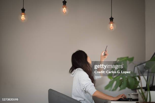 a young asian woman adjusts the power of the ceiling led lights from her mobile phone while working from home on her desktop computer sitting on an office chair in the living room - reportage home stock pictures, royalty-free photos & images