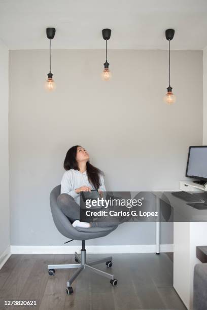 a young asian woman adjusts the power of the ceiling led lights from her digital tablet while working from home on her desktop computer sitting on an office chair in the living room - reportage home stock pictures, royalty-free photos & images