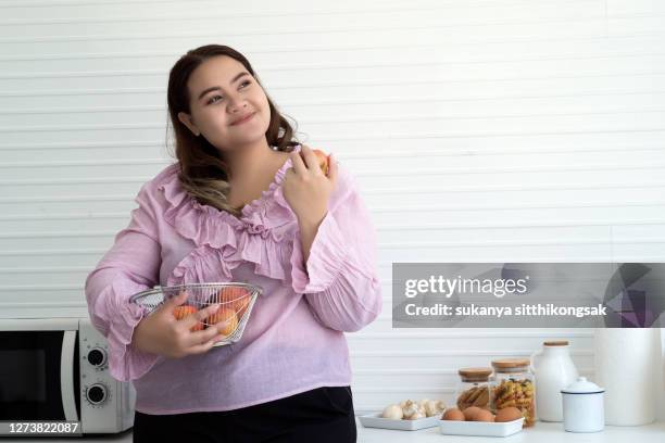 overweight woman  eating an apple in her kitchen at home for weightloss in her kitchen at home. - fat woman stock-fotos und bilder