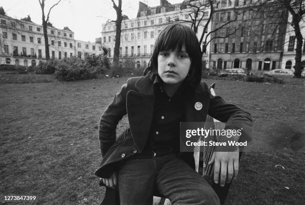 British songwriter, musician, and record producer Ricky Wilde wearing an overcoat a Smiley badge on the lapel, as he poses sitting on the arm of a...