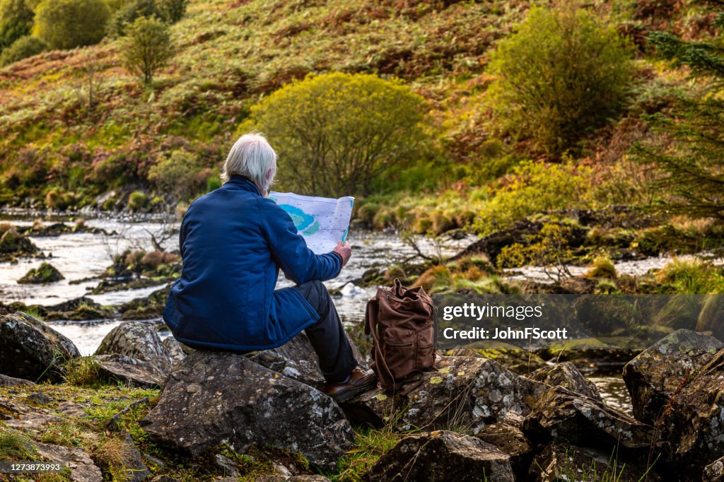 Senior man looking at a map while sitting on a rock beside a Scottish river in rural south west Scotland.