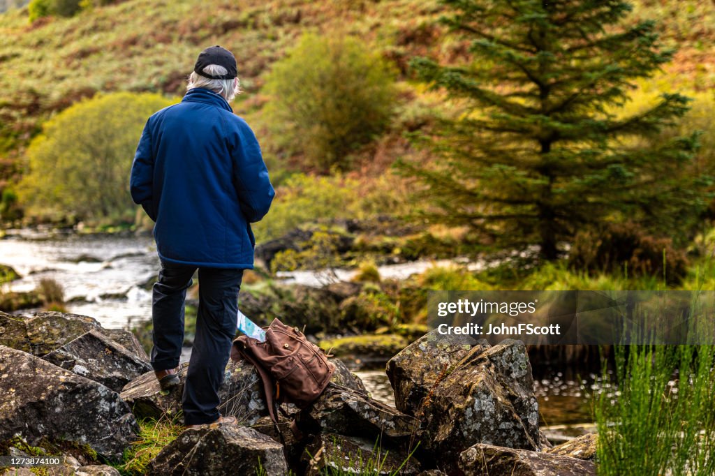 退休男子獨自站在蘇格蘭西南部農村一條蘇格蘭河旁的岩石上。