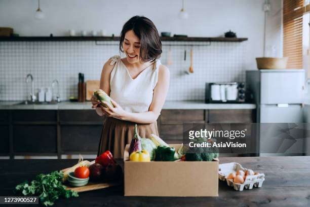 hermosa joven asiática sonriente recibió una caja llena de alimentos orgánicos coloridos y frescos ordenados en línea por el servicio de entrega a domicilio. ella está ordenando los comestibles y preparándose para cocinar una comida saludable - ecosistema fotografías e imágenes de stock
