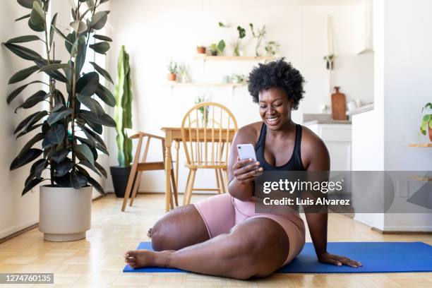woman using phone after exercising at home - curvy black women stock-fotos und bilder