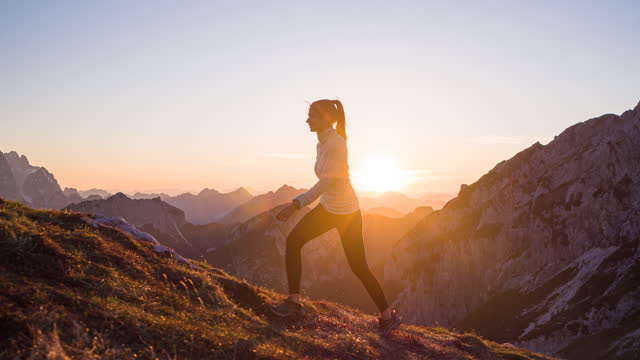 Fit woman athlete maintaining a healthy lifestyle, hiking in mountains over rocky trails and grassy slopes at sunset