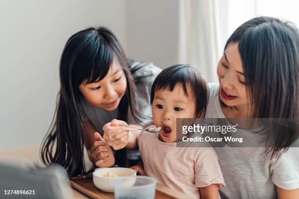 young girl helping her mother feeding food to toddler sister - asian spoon feeding stockfoto's en -beelden