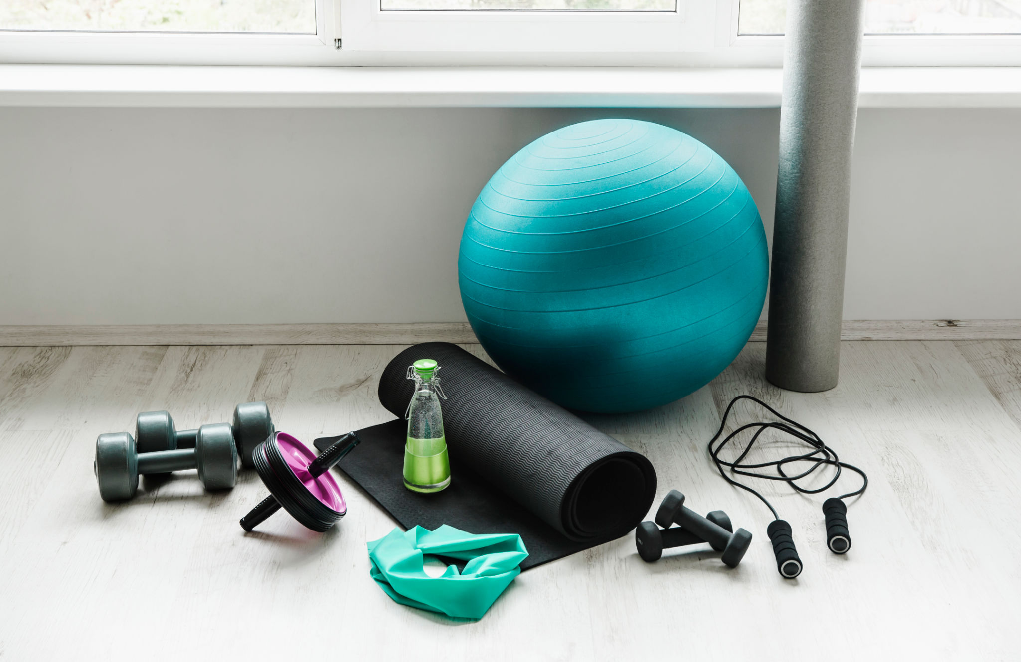 Group of different exercising equipment on white home gym floor. Fitness ball, round foam roller, resistance exercise latex band, jumping rope, dumbbells, yoga mat. Fit lifestyle concept. Group of different exercising equipment on white home gym floor. Fitness ball, round foam roller, resistance exercise latex band, jumping rope, dumbbells, yoga mat. Fit lifestyle concept.