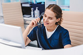 Young female receptionist talking on phone in clinic while sitting and looking on pc monitor