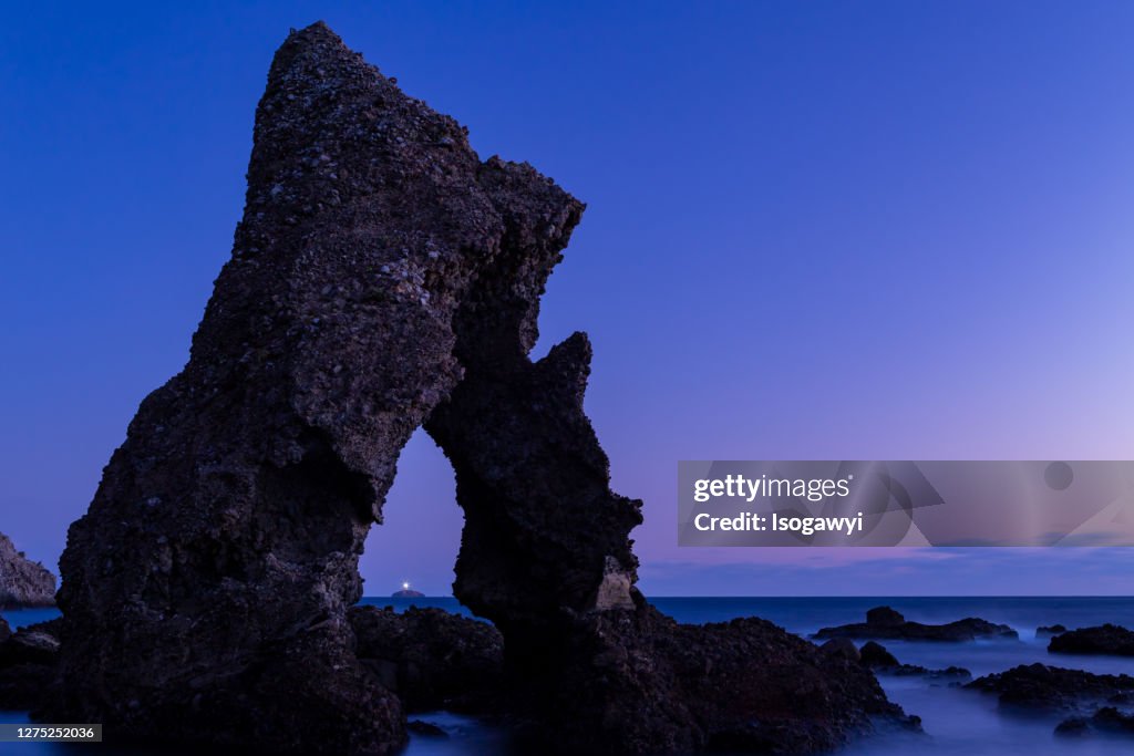 A Lighthouse On The Island Through The Bizarre Rock Against Sky During Sunset