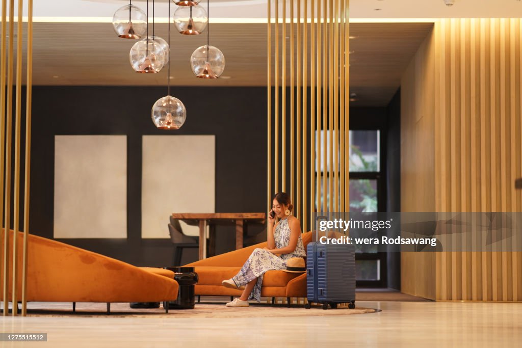 Young tourist woman sitting on sofa at hotel lobby lounge using phone to talking and smiling