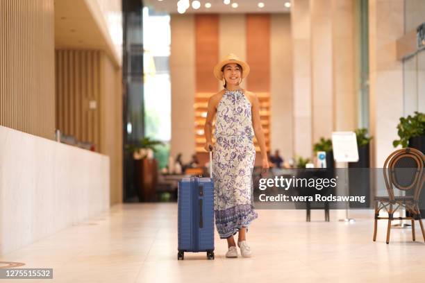 tourist woman with sun hat carrying wheeled luggage walking in front of the hotel reception desk while smiling and looking away - airport lounge luxury stockfoto's en -beelden