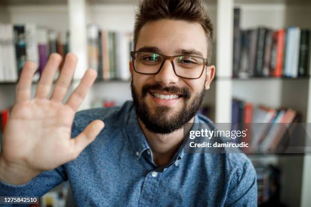 jonge mens die met hand op videovraag bij huisbureau golft - waving hand stockfoto's en -beelden