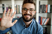 Young man waving with hand on video call at home office