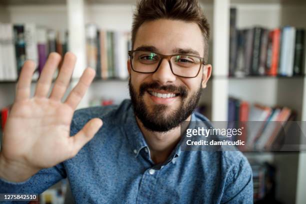 young man waving with hand on video call at home office - looking at camera stock pictures, royalty-free photos & images