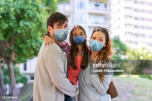 beautiful latin american couple carrying their daughter enjoying a day outside all wearing protective facemasks while looking at camera - argentina covid stock pictures, royalty-free photos & images