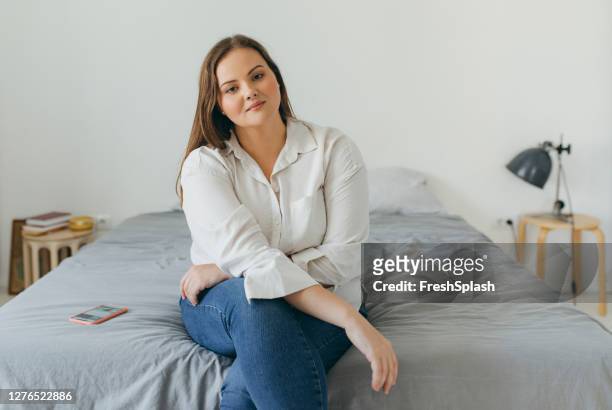 一個自信的加號女人坐在她的床上的肖像, 看著相機。 - curvy woman 個照片及圖片檔