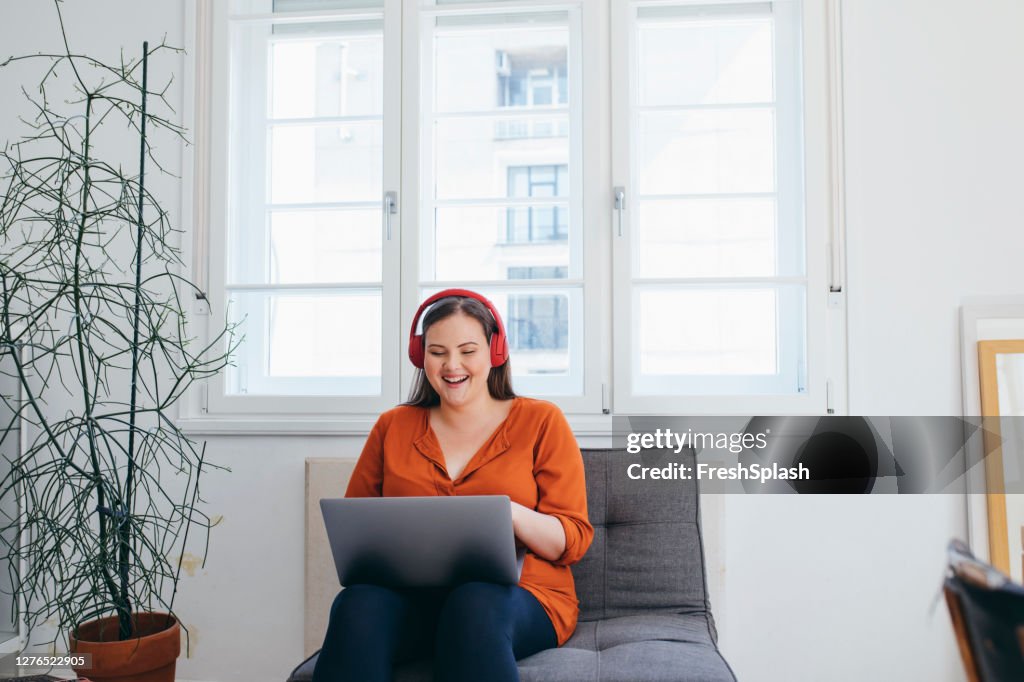 彼女のラップトップPC上で何か楽しいものを見てヘッドフォンを身に着けているハッピープラスサイズの女性