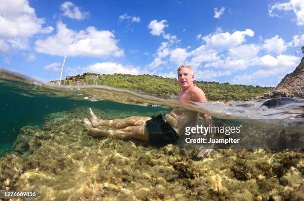 hombre joven disfruta de un dia de playa sentado sobre una roca bajo el agua en una cala de la isla de menorca - hombre sentado stock-fotos und bilder