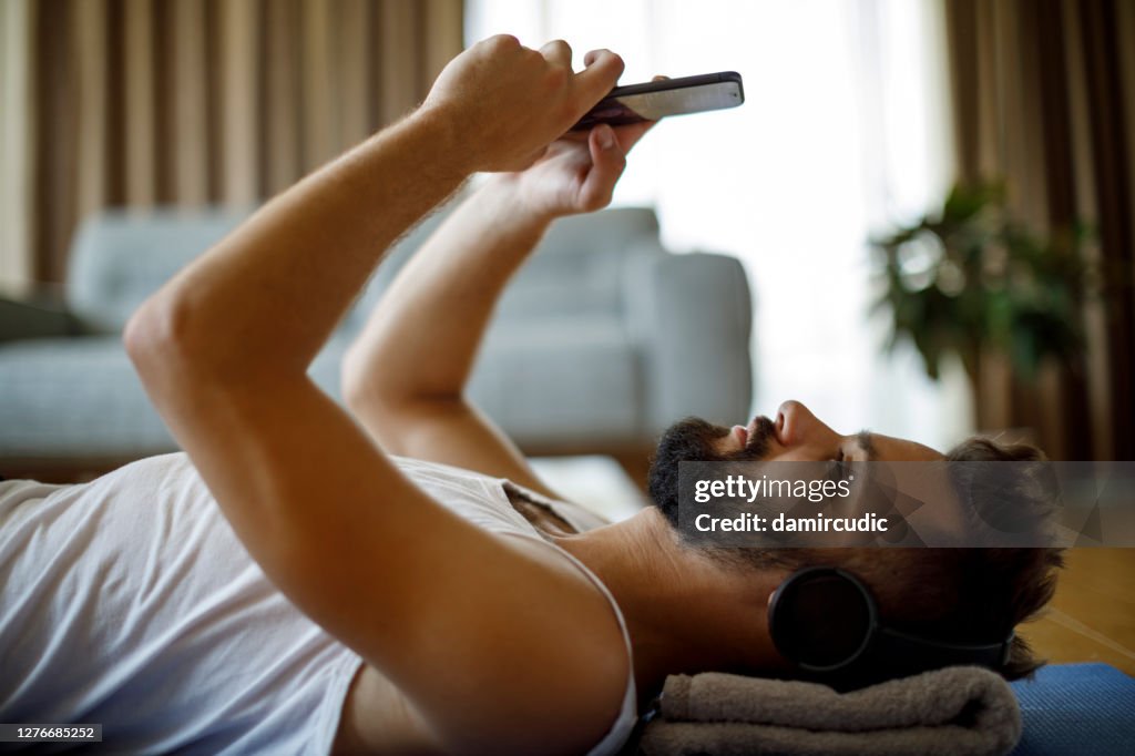 Young smiling man with headphones and mobile phone relaxing after exercising at home
