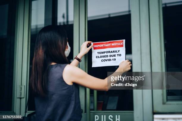 an asian woman small business owner with protective face mask putting on a closing down notice onto a store window due to the covid-19 pandemic - 閉店売り尽くしセール ストックフォトと画像