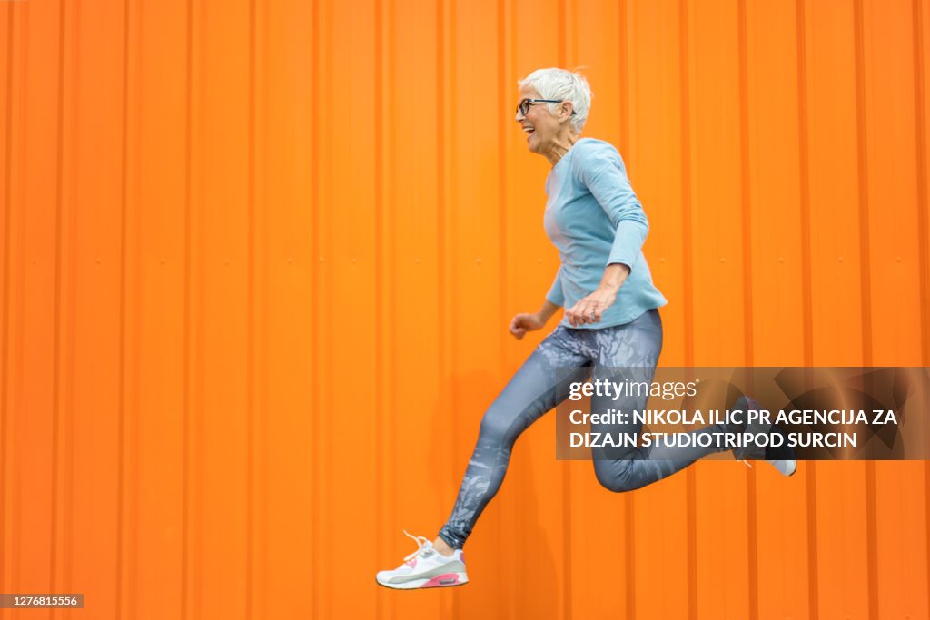 Glückliche Seniorin springt vor orangefarbenem Hintergrund.
