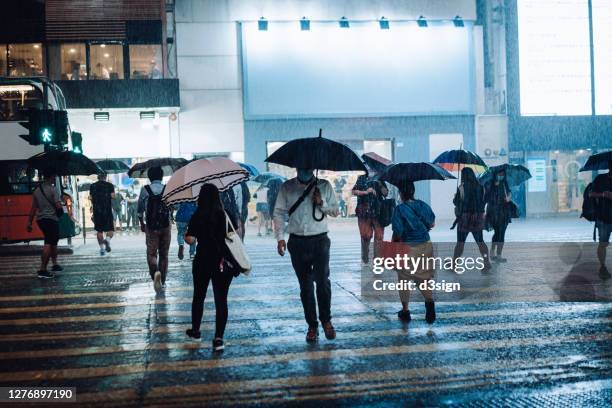 crowd of people with protective face mask carrying umbrellas crossing the street in downtown district in the city, against the reflection of glowing neon lights and city buildings in heavy rain at night - heavy rain ストックフォトと画像