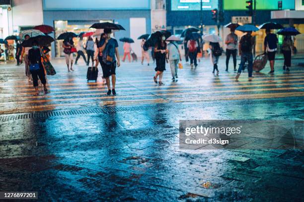 crowd of people with protective face mask carrying umbrellas crossing the street in downtown district in the city, against the reflection of glowing neon lights and city buildings in heavy rain at night - heavy rain stock-fotos und bilder